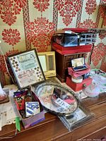 Photo of assorted items including framed Canadian currency, small vintage style mirror, metal framed mirrored trays, scissors, cutters, and red and pink jewelry boxes stacked on a side table with patterned wallpaper background.