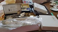 Photo showing assortment of silver utensils including spoons, knives, and mother of pearl utensils in boxes and plastic wrapping.