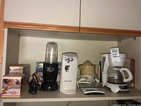 Shelf with Magic Bullet blender, Black & Decker can opener, Braun citrus juicer, Sunbeam coffee maker, digital scale, and boxed cheese slicer