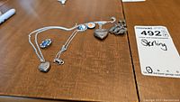 Two sterling silver necklaces on wooden surface; one features a heart-shaped locket, the other has a seahorse pendant, with a blue floral brooch visible.