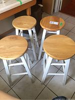 Four wooden stools with round natural wood seats and white painted legs standing on tiled floor, showing condition and design.