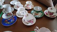 Top view showing all teacups and saucers arranged in a semi-circle on a table, displaying a variety of floral and fruit designs with gold trim and different base colors.