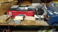 Photo of various items on a wooden bench including a red and black plastic toolbox, a blue metal toolbox, cardboard boxes, and miscellaneous tools and boxes.
