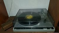 Side view of Technics SL-220 turntable with smoke-colored dust cover, vinyl record on platter, and wooden box beside it.