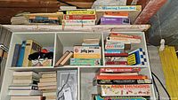 Overview photo showing collection of vintage board games and books inside a divided shelf with visible titles such as Scrabble, Password, Spirograph, Jelly Jars, and weaving loom set.
