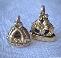 Two antique gold-tone fobs with intricate design and hanging loops, displayed on light fabric.