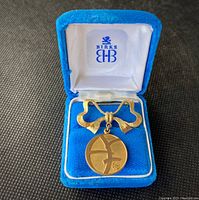 Front view of a Birks gold fill brooch with bow shaped top and circular pendant inside blue velvet display box with white silk lining and Birks logo.