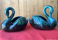 Pair of Blue Mountain Pottery swan figurines on a red surface against a wooden background, showing both swans side by side.