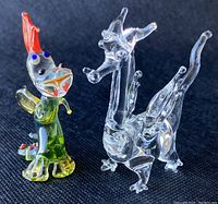 Photo showing both dragon figurines together on a dark textured background, one clear glass and one green with red and blue accents.