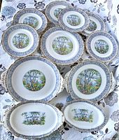 Overview of all 11 pieces on floral cloth background showing 6 plates and 5 serving dishes arranged loosely in two rows