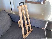 Wooden tabletop easel set on a gray couch, showing front three-quarter view with carrying handle on top.
