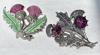 Pair of thistle brooches placed on white fabric; the first brooch is green and purple enameled with marcasite, the second is silver with purple stones and marcasite.