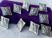 Ten silver square buttons with raised thistle design arranged on purple fabric, showing front faces.