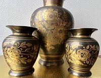 Three brass vases arranged upright, showing floral engraved pattern on round bodies.