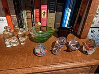 Front view showing all glass items arranged on a wooden surface with books in background including apothecary bottles, green glass dish, several glass animal figures, and paperweight.