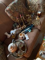 Overview of all items on a brown couch including brass candlesticks, candle holders, candles, brass bell, brass trivet, and tea set on brass tray.