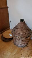 Green glass carboy bottle covered by large woven wicker basket with conical top placed on floor next to wooden round lazy susan tray.