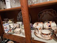 Wide shot showing the variety of teacups, saucers, and small plates arranged on a wooden shelf. Several floral designs and ornate patterns visible.