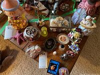 Overview showing vintage clown cookie jar, starfish decoration, glass bowl with decorative items, trinket box with flowers and ladybug pin, yellow rubber duck, whimsical brooch in blue box, and other assorted small figurines and keepsakes.