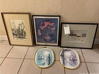 Five framed prints arranged on floor: three rectangular prints (cityscape, floral, cityscape with tram) and two oval fishing boat prints with gold frames.