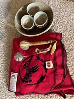 Overall view: red shawl, pottery bowl and cups, pewter pieces, and rattle