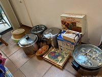 Wide view of all kitchen items grouped on floor showing pots, steamer, egg poacher, rice cooker, cheese board, and boxed apple parer.