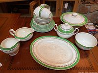 Full set displayed on table including cups, saucers, covered serving bowl, sugar bowl, cream pitcher, large serving platter and bowl all with green and floral border pattern.
