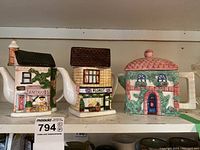 Three shop-shaped ceramic tea pots arranged side by side on a shelf showing detailed painted designs