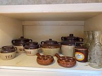 Full shelf showing ceramic canisters labeled Tea, Coffee, Sugar, small brown canister with label, escargot dishes, and clear glass decanters.