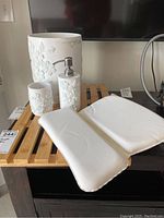 Set of white floral embossed ceramic cup, soap dispenser, waste basket, wood grate, and two white bath pillows on wood grate surface.