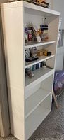 Front view of white laminate bookcase filled with various small items on three shelves and two empty shelves below.