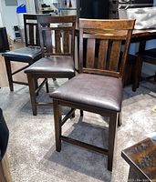 Four brown wooden counter chairs with padded brown faux leather seats arranged in a row on carpeted floor.
