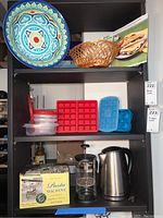 The kitchenware lot on shelves showing various items including a colorful ceramic plate, woven basket, plastic containers, silicone ice cube trays, French press, electric kettle, and pasta machine box.