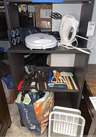 Shelf displaying a white electric hand mixer by Hamilton Beach with beaters, white sandwich maker or grill, blue star-shaped baking molds, various kitchen utensils including spatulas and brushes, and a cloth tote bag with storage bags on floor in front.