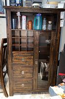 Front view of the dark wood wine cabinet showing 3 drawers on left, glass door with shelves on right, and cubby compartments above drawers.