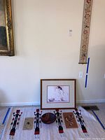 Full lot arrangement showing framed print on floor above area rug with Native American design, two art pieces on press board, carved bowl on rug, and hanging vertical tapestry.
