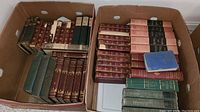 Two cardboard boxes filled with antique and vintage books with cloth bindings in red, green, pink, and blue colors.