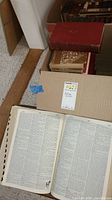 Webster's Third Dictionary open with visible text pages inside a cardboard box along with other antique books stacked above in the box.