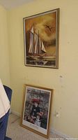 Photo showing both framed artworks leaning against the wall. One painting of a sailboat above, and a child portrait below.