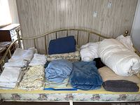 Overview of bed linens arranged on a yellow mattress including folded sheets, blankets, and comforters.