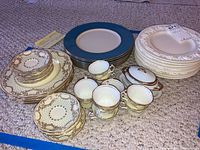 View of different patterned plates including those with intricate brown and gold floral designs, blue and gold rimmed plates, and white embossed plates, along with matching cup and saucer sets.