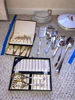 Wide view of lot showing boxed flatware sets (gold-toned and silver-toned), assorted silverware including spoons and ladles, and serving utensils arranged on carpet.