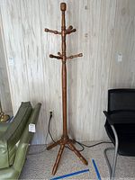 Full view of the wooden coat rack standing on carpeted floor against a light-colored wall, showing the four-legged base and multiple slotted pegs for hanging.