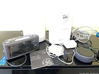 Photo showing GE digital alarm clock with power cord and manual, Amazon Echo Dot smart speaker, and power cables. All placed on black surface.