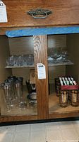 Cabinet shelf with clear water glasses, wine goblets, tall metal‐rimmed glasses, shot glasses, metal ice bucket, glass swizzle sticks, Georges Briard highball glasses