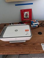 HP DeskJet 2755 printer on wooden surface with Canon photo paper and HP ink cartridges behind it