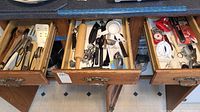 Photo of kitchen drawers open showing a variety of kitchen tools including cutting spoons, rolling pin, measuring spoons, kitchen timer, corkscrews, tongs and a cutlery sharpener in packaging.
