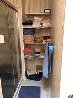 Shelving in bathroom showing multiple stacks of bath towels and washcloths in beige, blue, green, and rust colors along with some baskets and folded items near the floor.
