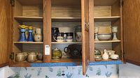 Wide view of a wooden cabinet interior with upper shelves holding multiple vintage pottery and mugs, including candlesticks, handled mugs, bowls, and other ceramic items