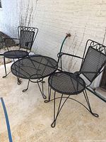Full view of the outdoor metal bistro set showing round table and two matching chairs on patio floor against white brick wall.